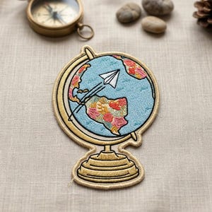 May include: Embroidered patch featuring a globe design with a paper airplane. The globe has a light blue background with floral patterns in pink, orange, and yellow. The patch has a gold border and base, and a compass is visible in the background.