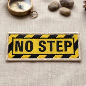 May include: A rectangular yellow patch with the words "NO STEP" in bold black letters. The patch has a black and yellow striped border, and a beige border. The patch is likely an iron-on or sew-on patch.