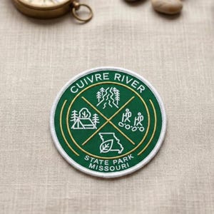 May include: A round, green patch with white trim featuring the text "CUIVRE RIVER STATE PARK MISSOURI". The patch has a gold border and includes illustrations of trees, a tent, a river, and people hiking. The patch is on a tan fabric.