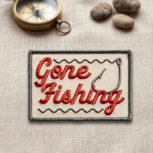 May include: Embroidered patch with the words "Gone Fishing" in red script. The patch features a fishing hook and a wavy line design. A vintage compass and small stones are in the background.