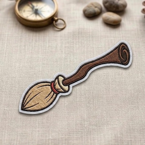 May include: Embroidered patch of a brown broom with a curved handle and a light tan bristle end. The patch has a white border and a red accent near the handle. The patch is designed to be ironed on or sewn onto fabric.