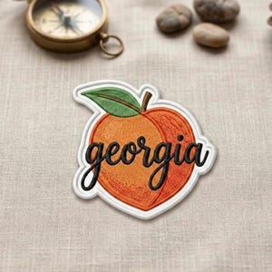 May include: Embroidered patch in the shape of a peach with the word "Georgia" in black cursive script. The peach is orange with a green leaf and a white border. The patch is on a beige fabric.