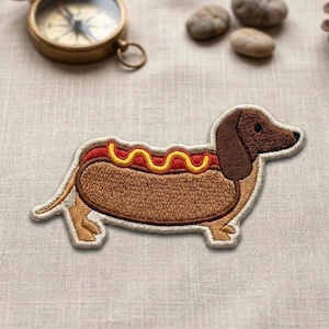 May include: Embroidered patch featuring a dachshund hot dog design. The patch has a brown body, a red and yellow topping, and a white border. The dog's head is brown with a black eye. The patch is likely for clothing or accessories.