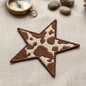 May include: A star-shaped patch with a brown and cream cowhide pattern. The patch is embroidered with a brown border and stitching. The patch is on a neutral linen surface, with a brass compass and small stones in the background.