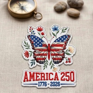 May include: Embroidered patch featuring a butterfly with wings designed as the American flag, surrounded by floral elements. The patch includes the text "AMERICA 250" and the dates "1776-2026". A vintage compass is visible in the background.