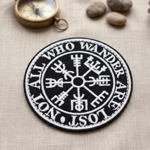 May include: A round black and white patch with the text "ALL WHO WANDER ARE NOT LOST" encircling a central symbol. The patch is embroidered with a white border. A vintage brass compass and small stones are in the background.