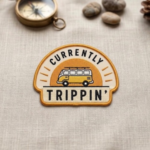 May include: Embroidered patch with the words "CURRENTLY TRIPPIN'" and a yellow van illustration. The patch has a cream-colored background with a yellow border and text. The patch is a travel-themed accessory.