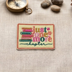May include: Embroidered patch with the words "just one more chapter" in pink, teal, and gold. The patch has a light yellow background and a red border. The patch is rectangular and features stacked book designs.