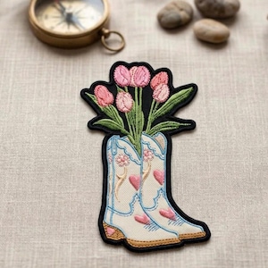 May include: Embroidered patch featuring a pair of white cowboy boots with pink hearts and floral details, holding pink tulips with green leaves. The patch has a black border and is set against a neutral background.