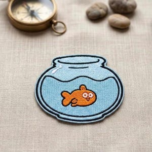 May include: Embroidered patch featuring a cartoon goldfish in a blue fishbowl. The fish is orange with large eyes, and the bowl has a black outline. The patch is on a neutral-colored fabric, suitable for clothing or accessories.