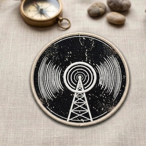 May include: Round black and white patch featuring a radio tower design. The patch has a distressed look with a white border. A vintage compass and small stones are in the background.