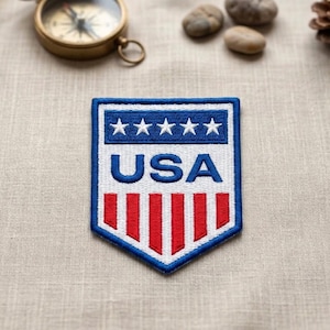 May include: Embroidered patch with a blue border, featuring the word "USA" in white block letters. Above the text are five white stars on a blue background. Below the text are red vertical stripes. The patch is on a tan fabric surface.