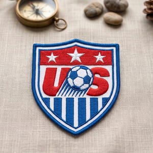 May include: A shield-shaped patch with a soccer ball and the letters "US". The patch has a red top with three white stars, a blue and white soccer ball, and blue and white stripes at the bottom. The letters "US" are in red.