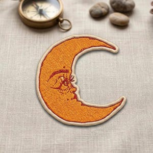 May include: Embroidered orange crescent moon patch with a face design. The patch features a cream-colored border and intricate details in red thread, perfect for adding to clothing or accessories.