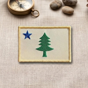 May include: A rectangular patch featuring the Maine state flag design. The patch has a cream-colored background with a blue star in the upper left corner and a green pine tree in the center. The patch is bordered with a gold-colored stitching.