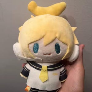 May include: A plush toy featuring a character with short yellow hair, a yellow bow on top, and teal embroidered eyes. The figure wears a white sailor-style shirt with a yellow tie, dark blue striped accents on the sleeves, and white headphones.