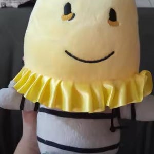May include: A soft toy with a yellow head and a smiling face. The toy has a yellow ruffled collar and a body with white and black stripes. The toy has small arms and legs.