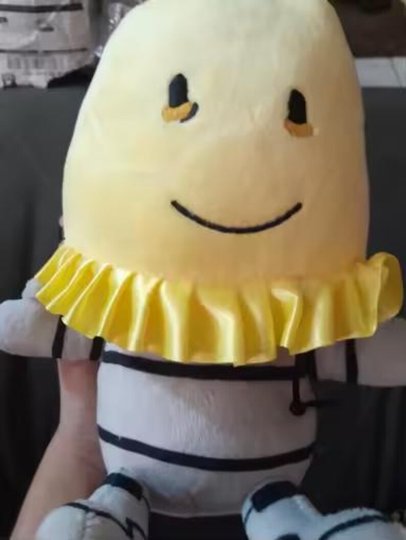 May include: A soft toy with a yellow head and a smiling face. The toy has a yellow ruffled collar and a body with white and black stripes. The toy has small arms and legs.
