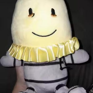 May include: A soft toy with a smiling face and a yellow ruffled collar. The toy has a light yellow head, grey and white striped body, and black accents. The toy is approximately 30 cm tall.