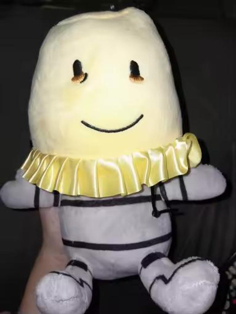 May include: A soft toy with a smiling face and a yellow ruffled collar. The toy has a light yellow head, grey and white striped body, and black accents. The toy is approximately 30 cm tall.