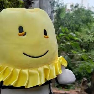 May include: A soft toy of a smiling yellow character with black eyes and a black smile. It features a yellow ruffled collar and a black and white striped body. The background is a blurred outdoor scene.