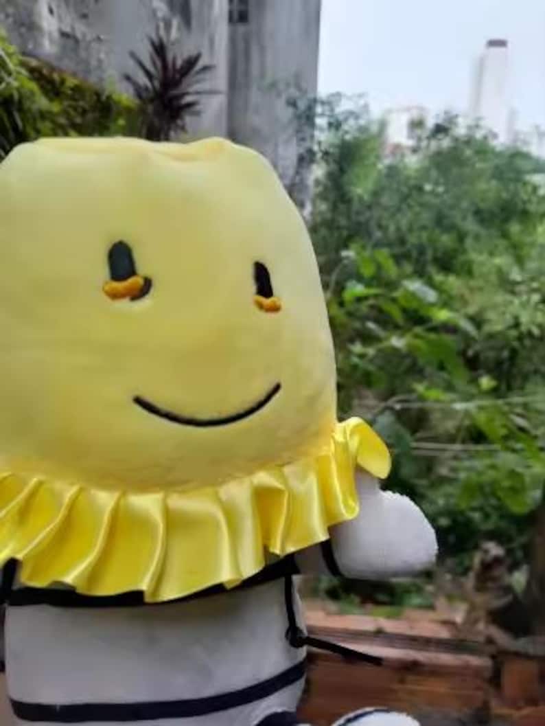 May include: A soft toy of a smiling yellow character with black eyes and a black smile. It features a yellow ruffled collar and a black and white striped body. The background is a blurred outdoor scene.
