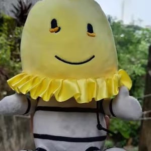 May include: A soft toy with a yellow head and a smiling face. The toy has a yellow ruffled collar, white body with black stripes, and white arms and feet. The toy is designed to look like a cartoon character.