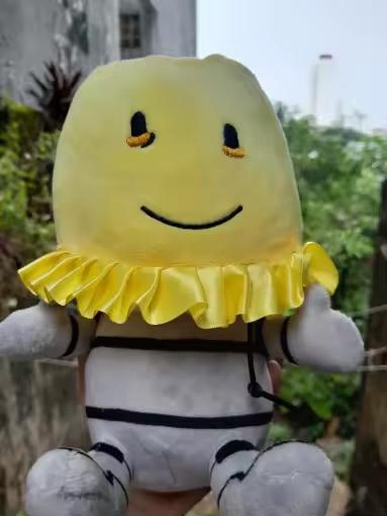 May include: A soft toy with a yellow head and a smiling face. The toy has a yellow ruffled collar, white body with black stripes, and white arms and feet. The toy is designed to look like a cartoon character.