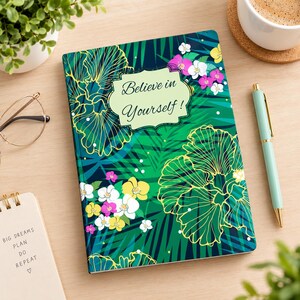 May include: A rectangular notebook with a dark green background featuring a pattern of tropical palm leaves and colorful orchid flowers in white, yellow, and pink. The cover displays the text Believe in Yourself inside a light green label.