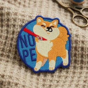 May include: A circular embroidered patch featuring a tan and white Shiba Inu dog on a blue background. The dog is wearing a red collar and leash, looking away with a stubborn expression. The text NOPE is embroidered in dark blue on the left side.