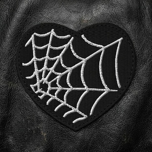 May include: A black heart-shaped patch with a white spiderweb design, stitched onto a black leather surface. The patch is a decorative element, possibly for clothing or accessories.