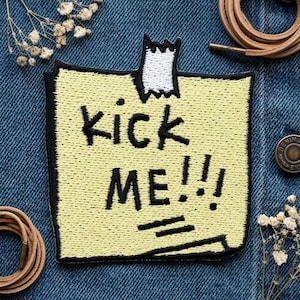 May include: A yellow patch with the words "kick me!!!" in black, resembling a sticky note, is affixed to a denim surface. The patch has a black border and is held in place with white tape. Decorative elements include leather cords and small white flowers.