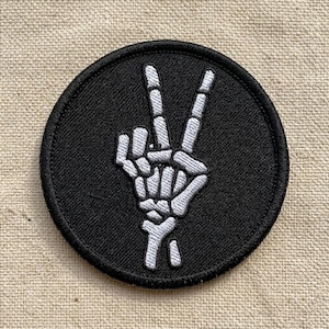 May include: A round, black patch featuring a white, skeletal hand making the peace sign. The patch is embroidered with detailed bone structures and has a black border. The background is a neutral, textured fabric.
