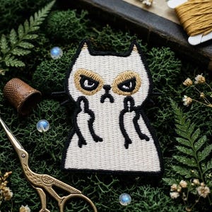 May include: An embroidered patch featuring a white cat with a grumpy expression, gold-colored fur around its eyes, and black outlines. The cat is depicted with its arms raised, showing its middle fingers in a defiant gesture against a dark background.
