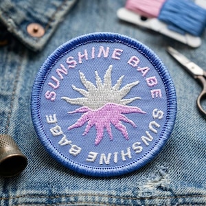 May include: A round, blue embroidered patch with the words "SUNSHINE BABE" in pink and white. The patch features a sun design in white and pink. The patch is on a denim jacket.
