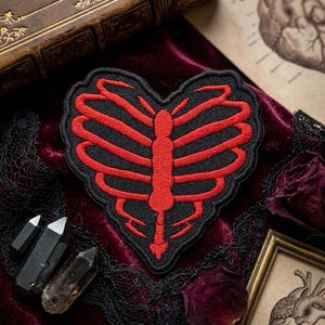 May include: A heart-shaped embroidered patch featuring a red rib cage design on a black background. The patch has a scalloped edge and depicts a stylized human thoracic cage structure with a central sternum and curved ribs forming a heart shape.
