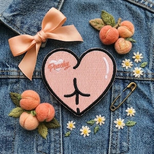 May include: A denim jacket adorned with a peach-colored bow, embroidered peach clusters, and a pink heart patch with the word "Peachy". Additional details include embroidered daisies and a gold safety pin.