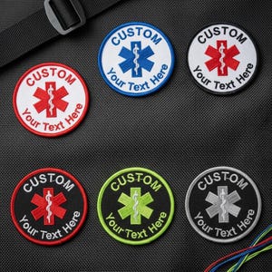 May include: Embroidered patches with a Star of Life symbol in various colors: red, blue, green, and silver. Each patch has the word "CUSTOM" and the text "Your Text Here" in a circular design. The patches are round and have a white border.