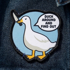 May include: Embroidered patch featuring a white duck with a yellow beak and feet, holding a knife. The duck is set against a light blue circle with the text "DUCK AROUND AND FIND OUT" in a speech bubble. The patch is on a denim background.