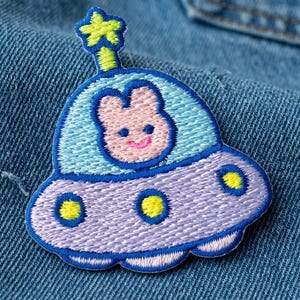 May include: Embroidered patch featuring a cartoon spaceship with a smiling pink bunny pilot. The spaceship is lavender with blue and yellow accents, and a star on top. The patch is sewn onto a denim background.