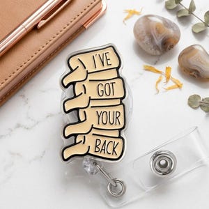 May include: A badge reel featuring a cream-colored acrylic charm shaped like a stack of four human vertebrae. The charm has black outlines and black text that reads I'VE GOT YOUR BACK. The badge reel includes a clear plastic strap and a metal snap.