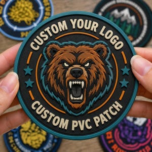 May include: A circular PVC patch featuring a central illustration of a snarling brown bear with bared teeth. The text CUSTOM YOUR LOGO and CUSTOM PVC PATCH is embossed in beige lettering along the black border, accented by teal stars and rings.