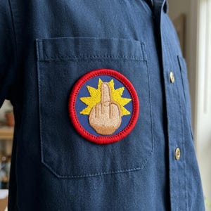 May include: A navy blue shirt with a round embroidered patch on the pocket. The patch features a hand giving the middle finger, surrounded by a yellow burst design and a red border. The shirt has gold-colored buttons.