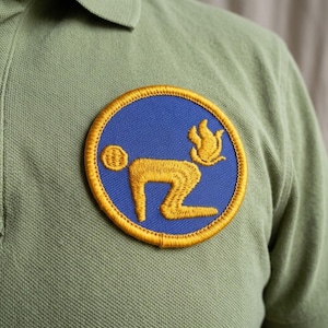 May include: A round embroidered patch with a blue background and a gold border. The patch features a stylized gold figure in a kneeling position, with a flame and a small circle.