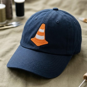 May include: Navy blue baseball cap with an embroidered orange and white traffic cone patch. The cap has a curved brim and a small button on top. The hat is made of a soft, durable fabric.