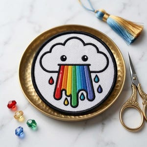 May include: A circular embroidered patch featuring a white cloud with a black outline and two black eyes. A vibrant rainbow drips from the cloud, accompanied by small, colorful raindrops in red, blue, green, and purple, all set against a white background.