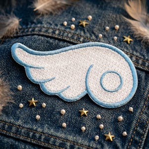 May include: An embroidered patch shaped like a stylized wing with white thread and a light blue border. The wing features a circular detail on the right side and is placed on a dark blue denim fabric decorated with small gold stars and white beads.