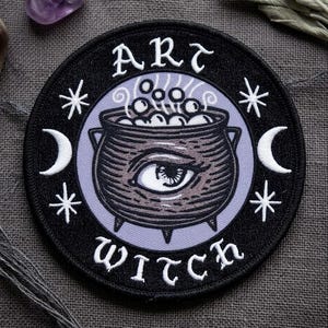 May include: A round black patch with the words "ART WITCH" in white script. The patch features a cauldron with an eye inside, surrounded by stars and crescent moons. The cauldron is filled with bubbles, and the background is a light purple.