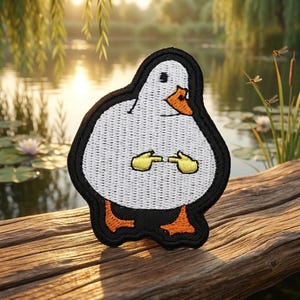 May include: An embroidered patch featuring a white duck with an orange beak and orange feet. The duck is depicted with a rounded body, black outline, and yellow hands positioned in front of its chest with index fingers pointing toward each other.