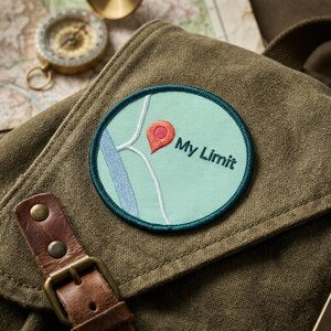 May include: A round embroidered patch with a map design and the words "My Limit." The patch features a light blue background with a teal border, a red location marker, and a blue river. The patch is on a green canvas bag.